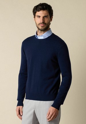 Marineblauwe crewneck trui van zacht materiaal met geribbelde manchetten en zoom, gedragen over een lichtblauw overhemd met kraag, gecombineerd met lichte broeken.