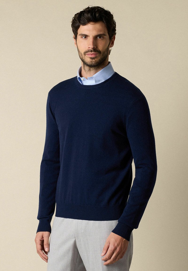 Námořnický svetr s kulatým výstřihem vyrobený z měkkého materiálu s žebrovanými manžetami a spodním lemem, nošený přes světlou modrou košíčkovou košili, spárovaný se světlými kalhotami.