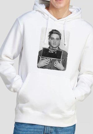 Person trägt weißen Hoodie und hält mit den Händen in den Taschen ein Namensschild mit der Aufschrift "Presley Elvis A." sowie eine schwarz-weiße Mugshot-Fotografie von Elvis Presley.
