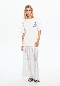 T-shirt en coton blanc avec un graphique bleu sur la poitrine gauche, associé à une jupe maxi blanche en dentelle avec des rayures verticales et des couches transparentes.