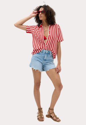 Kvinde iført rød og hvidstribet skjorte, rød bikinitop, denimshorts, røde solbriller og brune remsandaler, poserer med den ene hånd på solbrillerne.