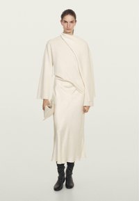 Crème-kleurige gebreide jurk met een draperend ontwerp, lange mouwen en een ontspannen silhouet. Gecombineerd met zwarte kniehoge laarzen en een clutch.