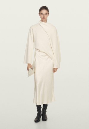 Cream-colored knit dress with a draped design, long sleeves, and a relaxed silhouette. Paired with black knee-high boots and a clutch.