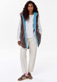 Hnědá bezrukávová vesta s nadýchanou texturou, tyrkysovým podšívkou, zipovým zapínáním a růžovými akcenty, nošená přes bílý svetr a světlé kalhoty.
