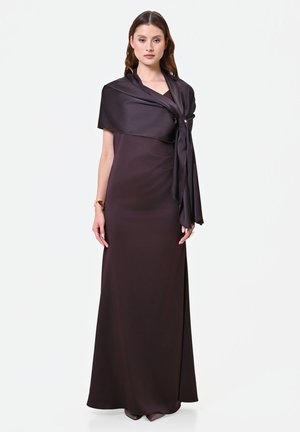 Femme portant une longue robe marron foncé avec un grand châle assorti noué à l'épaule, posant devant un fond blanc uni.
