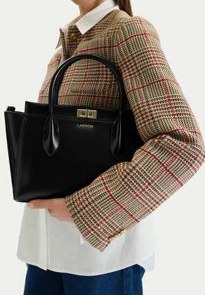 Donna che indossa una giacca a quadri beige e rossa tiene una borsa nera Lasocki con dettagli dorati e doppi manici.