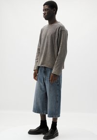 Maglia grigia a maniche lunghe, leggermente oversize con bordi grezzi, abbinata a pantaloni shorts in denim blu oversize. Scarpe e calze nere completano l'outfit.