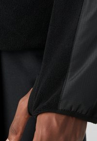 Svart fleecejakke med teksturert stoff på kroppen og glatte, blanke svarte ermer. Ermene har en trimdesign for god passform.