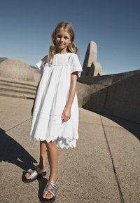 Hvid kortærmet kjole med indsnøring ved taljen, blonderkant ved kanten og en løs pasform, kombineret med sølvsandaler med brede remme og sort sål.