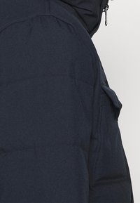 Marinblå puffjacka med mjuk textur, med dragkedjeslutning, stora sidofickor och sydda paneler för isolering.