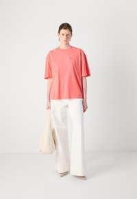 Coral cotton T-shirt with short sleeves and a rounded neckline, paired with white flared trousers and a beige bag. Simple, minimal design.
