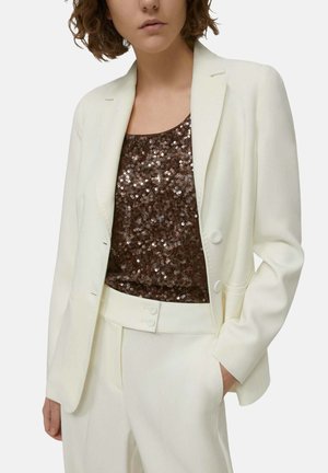 Blazer bianco su misura in tessuto liscio, indossato sopra una maglia marrone con paillettes. L'outfit include pantaloni crema coordinati con dettagli a bottoni.