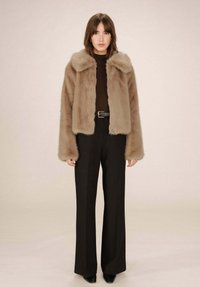 Giacca in pelliccia sintetica di un morbido beige con un ampio colletto, abbinata a pantaloni neri a vita alta e scarpe nere a punta, in un contesto neutro.
