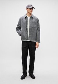 Grau wolles Hemdjacke mit einer Vordertasche, getragen über einem weißen T-Shirt, kombiniert mit dunklen Jeans und schwarzen Schuhen, abgerundet mit einer grauen Mütze.