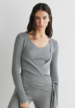 Frau mit dunklem Haar, die ein eng anliegendes, geripptes, graues Strick-Wickeloberteil trägt, das an der Taille gebunden ist, und passende gerippte Strickhosen, stehend vor einer weißen Wand.