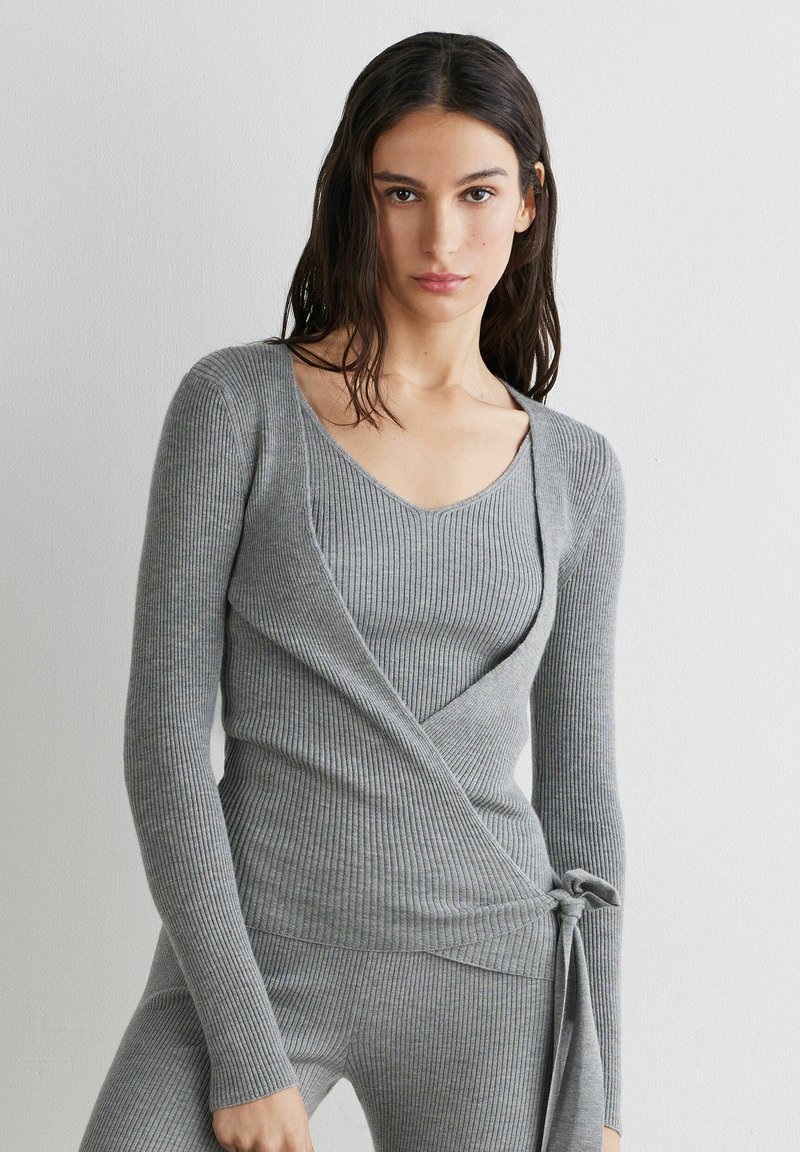 Femme aux cheveux foncés portant un haut cache-cœur gris côtelé ajusté noué à la taille et un pantalon assorti en maille côtelée, debout contre un mur blanc.