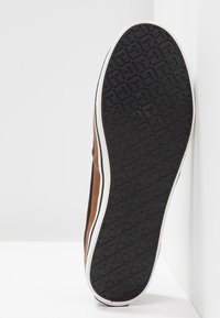 Suola di una scarpa casual con un motivo di battistrada in gomma nera che presenta forme a "T" ripetute, con intersuola bianca e tomaia in pelle marrone visibile lungo i bordi.