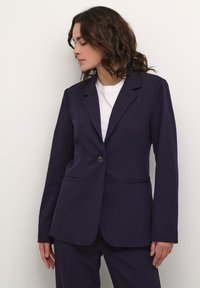Námořnický blazer vyrobený z hladké tkaniny, s jedním knoflíkovým zapínáním, klopami a dvěma předními kapsami, nošený přes bílý top.