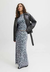 Donna che indossa un lungo vestito grigio con stampa leopardata, giacca di pelle nera drappeggiata sulle spalle, in piedi davanti a uno sfondo grigio chiaro uniforme.