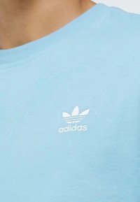 Světle modré bavlněné tričko s bílou vyšitou značkou Adidas a grafikou tří pruhů nad textem, kulatým výstřihem a krátkými rukávy.