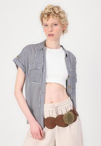 Chemise à rayures bleu marine et blanche, à manches courtes, avec deux poches. Assortie à un pantalon beige raccourci et une ceinture marron avec des accents circulaires.