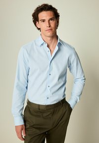Cămașă lungă cu mânecă, de culoare albastru deschis, cu guler cu nasturi, design ajustat și nasturi gri, asociată cu pantaloni de culoare măsliniu închis. Textură de țesătură netedă.