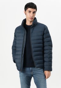 Navy puffer jacket with quilted design, high collar, full zipper, and side pockets. Paired with a knitted navy sweater and denim jeans.