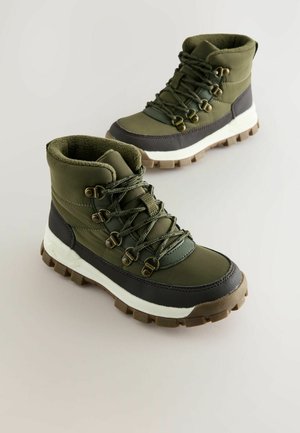 Ein Paar olivgrüne und braune isolierte Wanderstiefel mit dicken Gummisohlen und Metallösen auf einem hellgrauen Hintergrund.