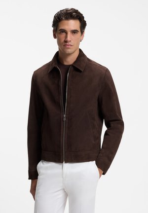 Mann trägt eine dunkelbraune Wildlederjacke mit Reißverschluss über einem dunklen Hemd und weißen Hosen, steht vor einem schlichten weißen Hintergrund.