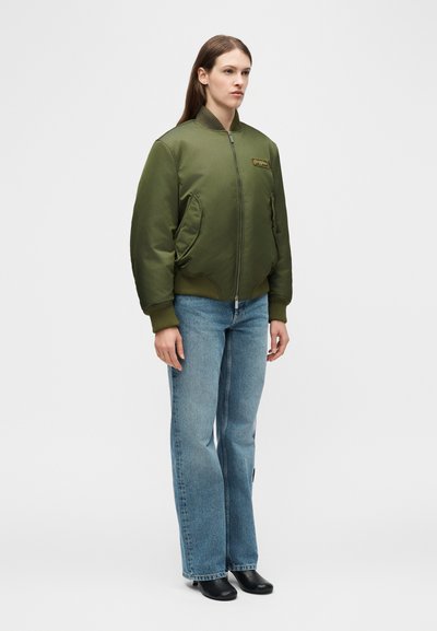 Γυναίκα που φοράει ανοιχτοπράσινο μπουφάν bomber με ριπ μανσέτες, μπλε τζιν με ίσια γραμμή και μαύρες μποτάκι αστραγάλου, στέκεται μπροστά σε απλό φόντο.