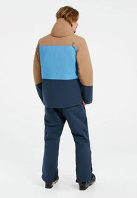 Chaqueta con bloques de color en marrón, azul y marino, que cuenta con una capucha, textura suave y un logo en la parte posterior, combinada con un pantalón marino a juego.