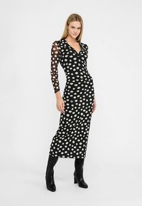 Zwarte jurk met witte polka dots, voorzien van lange doorschijnende mouwen, een V-halslijn en een nauwsluitend silhouet tot halverwege de kuit, gecombineerd met kniehoge zwarte laarzen.