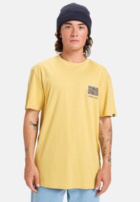 Žluté bavlněné tričko s kulatým výstřihem a krátkými rukávy; má grafický design kapsy a malou logo etiketu na lemu.