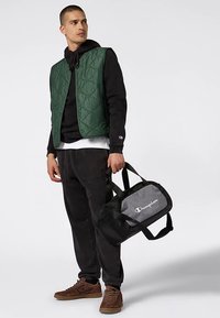 Gilet trapuntato verde sopra una felpa nera con cappuccio, abbinato a pantaloni della tuta neri e sneaker marroni. Borsa da viaggio Champion grigia e nera con logo.