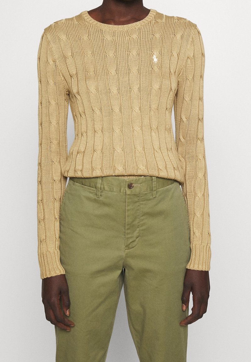 Personne portant un pull beige à torsades et un pantalon taille haute vert olive, debout avec les bras détendus le long du corps.