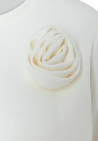 Valkoinen lyhythihainen yläosa, jossa on teksturoitu, kerroksellinen kangasruusukoriste saman sävyn eri vivahteissa sileää taustaa vasten.