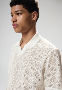 Polo en maille écru, arborant un motif en losanges ajouré. Manches courtes et col classique complètent le look.