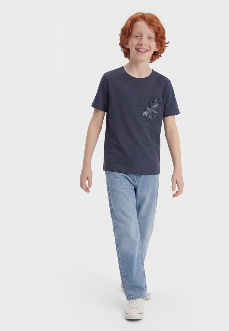 Smiling red-haired boy walks forward wearing a navy blue t-shirt with a floral pocket design, light blue jeans, and white sneakers.
