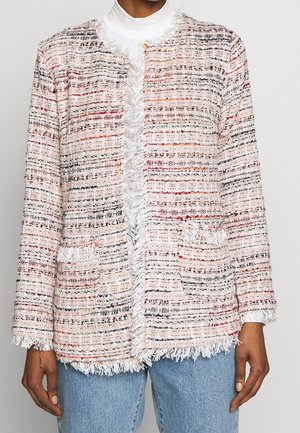 Kobieta ubrana w wielokolorową kurtkę z tweedu z frędzlami na krawędziach, do tego jasnoniebieskie jeansy i biały top z wysokim kołnierzem.