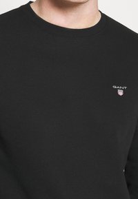 Felpa nera a girocollo realizzata in morbido tessuto, con un piccolo logo bianco sul petto. Design semplice con collo e orlo a costine.