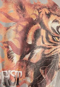 Koszulka z grafiką przedstawiającą szczegółową, ognistą ilustrację tygrysa w odcieniach pomarańczu i brązu, umieszczoną na jasnoszarym tle.