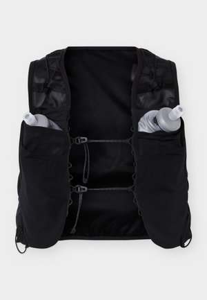 Junger Mann trägt schwarze Hydrationsweste mit zwei Wasserflaschen über einem dunklen Langarmshirt und grünen Cargoshorts, steht frontseitig.