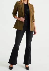 Olivově zelený blazer s figurálním střihem, doplněný černým topem s texturovaným vzorem a černými vysokými rozšířenými džíny. Černé podpatky.