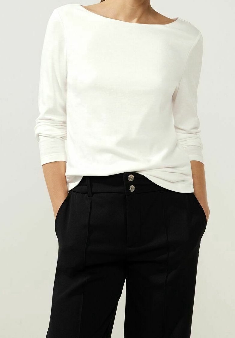 Person in einem weißen Langarm-Oberteil mit weitem Ausschnitt und schwarzen hochgeschnittenen Hosen mit zwei sichtbaren Knöpfen, die Hände in den Taschen.