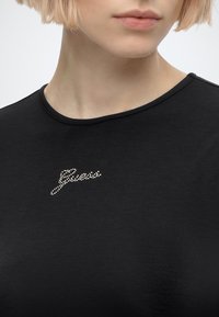 Gros plan d'une personne portant une chemise noire avec un petit logo scintillant "Guess" sur la poitrine, montrant les lèvres, le menton et le cou.