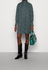 Robe à motif floral en bleu sarcelle foncé avec un col haut, des manches longues, accompagnée de bottes noires brillantes jusqu'au genou et d'un sac à main vert.