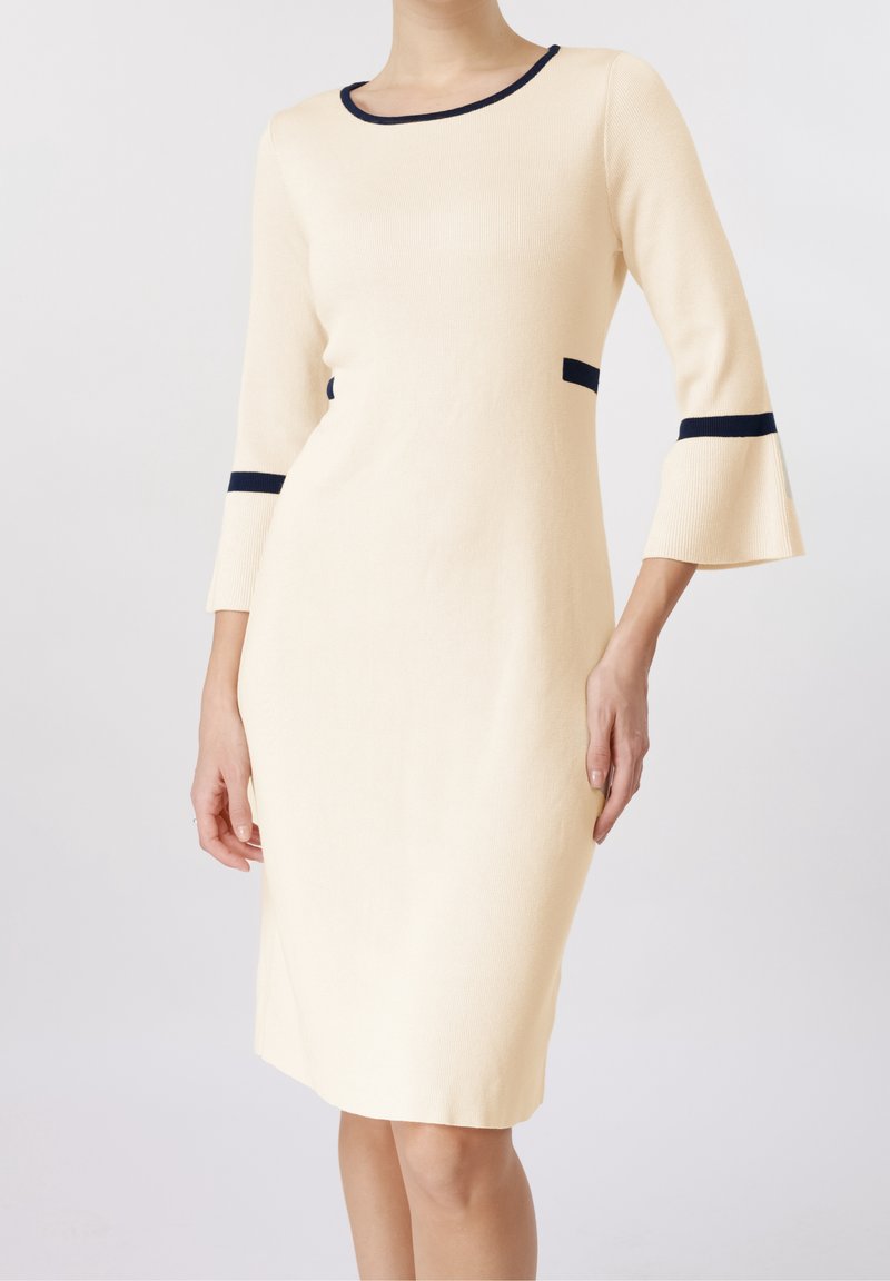 Femme portant une robe crème longueur genoux avec manches trois-quarts évasées et bordure marine à l'encolure, aux poignets et à la taille.