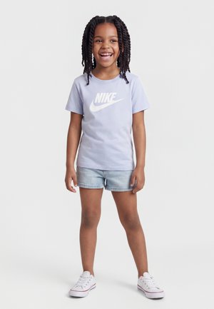 Mosolygós lány fonott hajjal, világoslila Nike pólót, farmer rövidnadrágot és fehér tornacipőt visel, egyszínű háttér előtt áll.