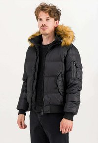 Giacca a vento nera con cappuccio foderato in pelliccia sintetica, chiusure con zip e bottoni a pressione, design trapuntato e polsini a coste, indossata sopra un maglione nero.