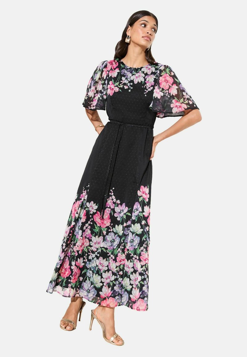 Vrouw in een zwarte maxi-jurk met kleurrijke bloemenprint op mouwen en zoom, gedragen met gouden sandalen met hoge hakken en opvallende oorbellen.